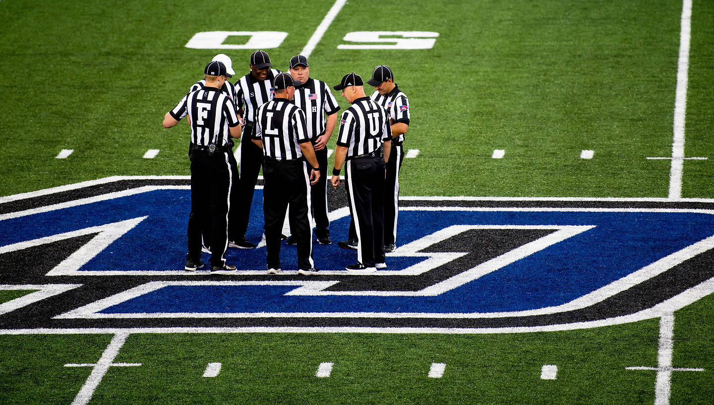 A group of football referees in black-and-white striped uniforms gather on a green field near a large blue and black logo.